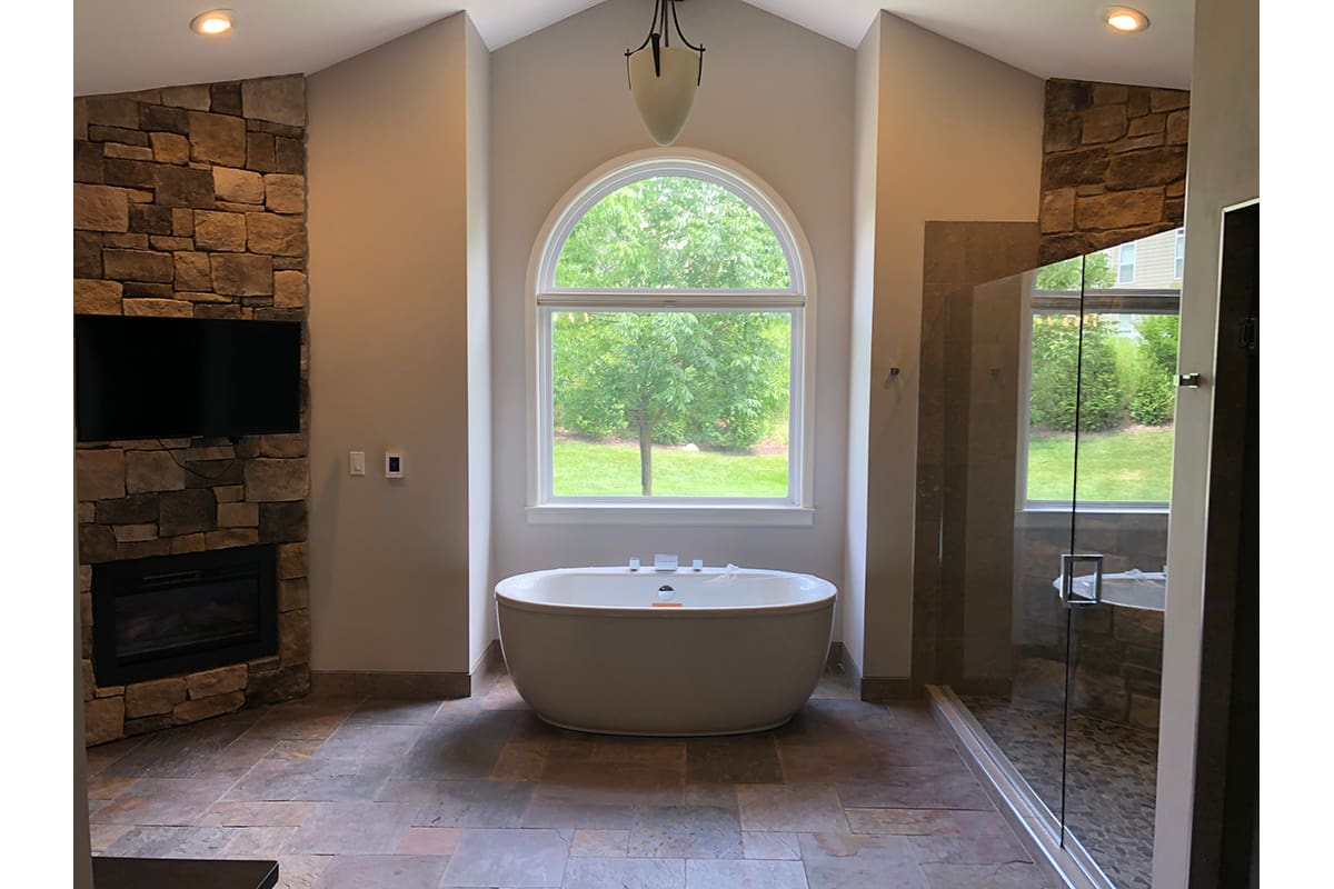 A large master bathroom featuring a freestanding tub and a pendant light. A window is centered in the room and opens up to a green tree.